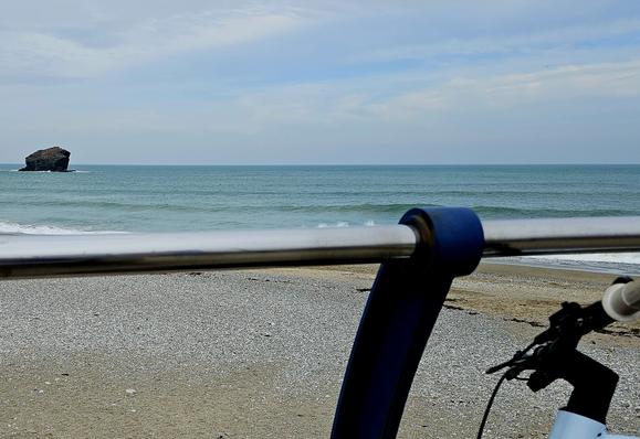A bike leans against the promenade railings at the beach. The sand is deserted, and there is a lone, black rock in the sea.