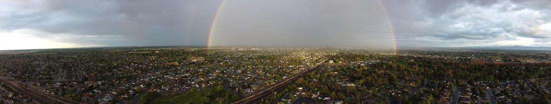 A wide pantographic image of a town in California's Central Valley. The skies are cloudy and there is a rainbow with light at the center.