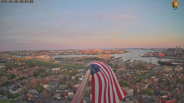 View from one of the National Park Service web cameras in Boston National Historical Park, looking over Boston Harbor from the top of the Bunker Hill Monument. In the foreground, a flag lit by the setting sun.