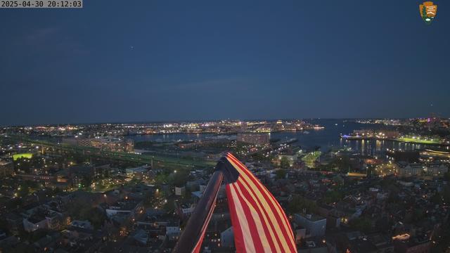 A second view from the National Park Service web camera in Boston National Historical Park, taken about a half a hour later. The image shows Boston Harbor from the top of the Bunker Hill Monument. In the foreground, a flag lit by the setting sun.