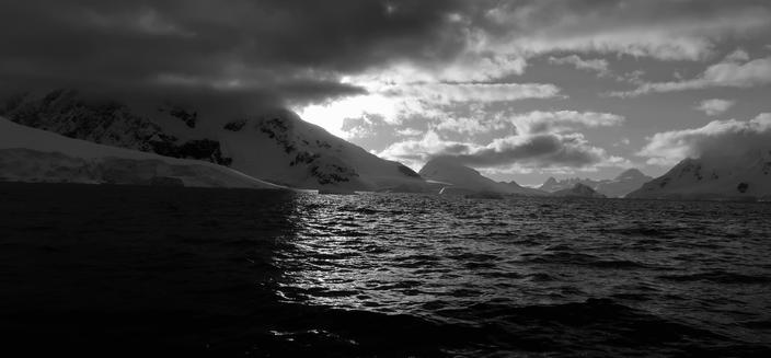 Black and white image half taken up by a calm sea with mountains in shadow either side, in the far distance are tall mountains entirely covered in snow