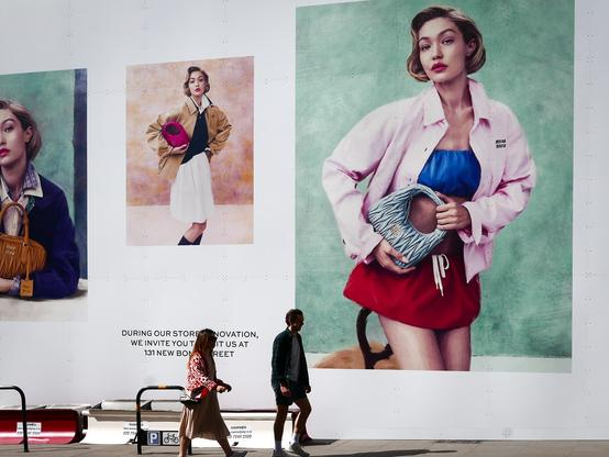 A man and a woman walking past Miu Miu in London as it is undergoing renovation. The shop front is covered with a huge white hoarding on which are three large photos of women wearing fashionable clothes and accessories. The photos dwarf the people walking past, especially the one at the right which is about twenty feet high featuring a woman who must be over twenty-five feet high.