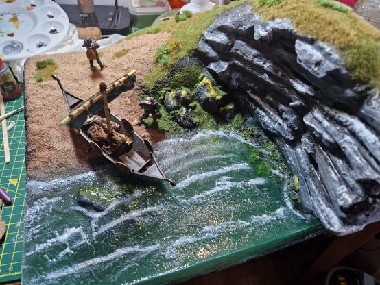 A view of a diorama from above and the front. It features a large cliff to one side, covered in a mixture of green and brown, rough grass. This leads down to a sandy beach with a figure on it carrying a sack away from a small drakkar (Norse longboat) that has been landed. The boat is full of sacks and ropes, and has a sail on a mast that has been handmade, furled up, and is lashed to the yard. The water is resin but has gentle white waves on its surface. The foot of the cliffs are green with seaweed and several larger boulders, topped with green-yellow lichen. The diorama scale is 28mm.
