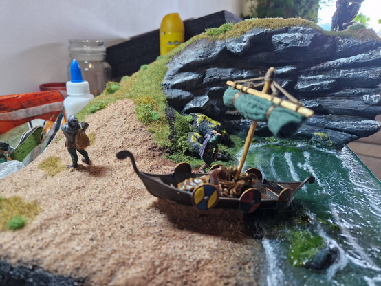 A view of the previous diorama from the side, looking down the beach towards the cliff, with a close-up of the drakkar. The sides of the drakkar are hung with hand-painted shields. In the background, there is modelling gear on the desk the diorama sits on.