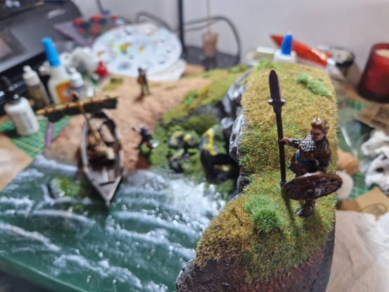 A view of the previous diorama from the cliff-top, with the beach out of focus in the background. The top of the cliff features a previously unseen figure; a shieldmaiden looking out to sea. In her right hand she holds an outrageously out-of-scale spear with knotwork on the tip. In her left hand she holds a round shield, painted with an anthropomorphic animal. Her eyes are heavily shadowed and her plaited blonde hair is loosely reminiscent of Lagertha, as played by Kathryn Winnick in the Vikings TV series. Or is meant to be.