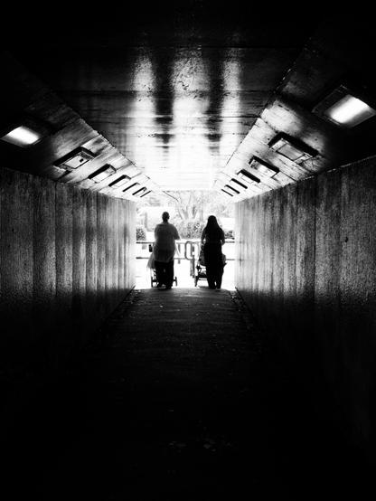 Two young women push prams into sunlight from the darkness of an underpass. The foreground is in dark shadow whilst the roof of the tunnel is bright with the distorted reflection of the two figures.