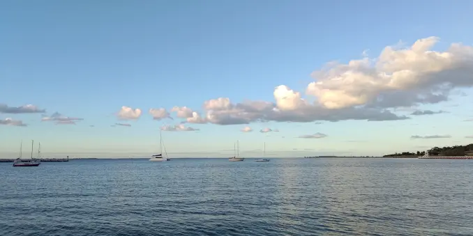 An outdoor photo towards the end of daylight, looking out towards a gentle sea that stretches more or less all the way to the horizon. A few small boats are visible in the distance. Some fluffy clouds dot the sky, partially lit by the sun.