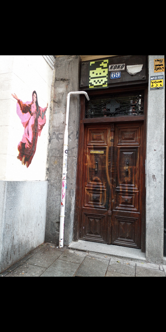 Photo of a door with cleaned off graffiti that has changed the colour of the wood stain, and street art above and to the side of it. There's a holy looking female figure floating and a pacman. The number is 69