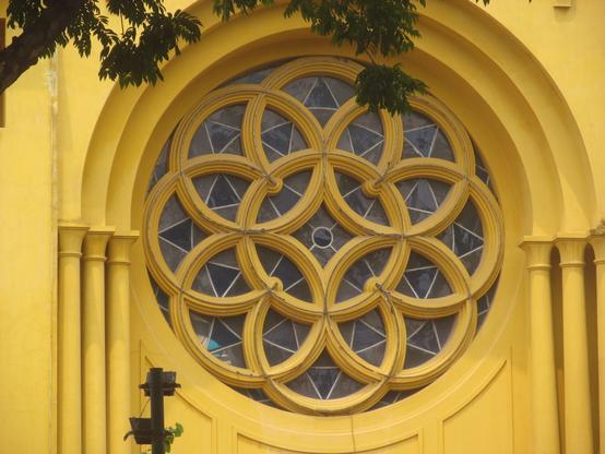 A photograph of a circular window of a church exterior. A central circle is overlapped by eight circles: the windows in the gaps have trianglar shapes. 
The church’s walls are yellow and part of a tree is visible.
