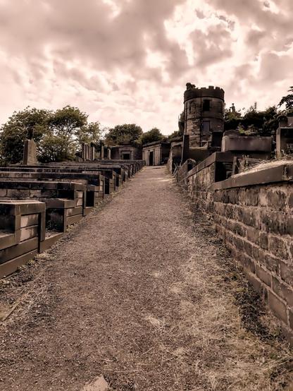 The image depicts a pathway through a cemetery, characterized by rows of aged and weathered gravestones and mausoleums on either side. The pathway is lined with stone and brick structures, some of which feature architectural elements like battlements, suggesting a historical or gothic style. The sepia tone of the image enhances its old-time feel, making the cemetery appear more solemn and possibly abandoned. Above, the sky is partly cloudy, adding to the atmospheric and perhaps melancholic mood of the setting.