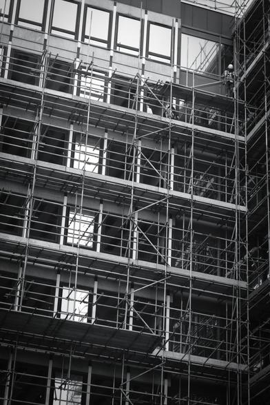 Black and white. A builder is standing on a wooden platform on scaffolding near the top of a building, looking down.