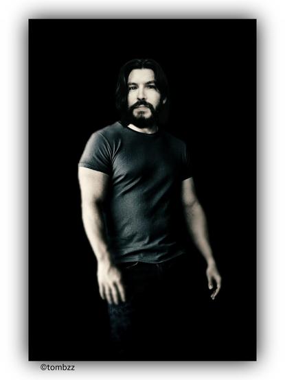 Portrait of a man against a black background. He is wearing a dark gray short-sleeved t-shirt that fits snugly, highlighting the muscular build of his arms and chest. His lower body is dressed in dark trousers. He stands upright with his arms relaxed at his sides.  

His face is sharp and expressive, with a well-defined jawline and high cheekbones. He has dark, thick eyebrows and deep-set eyes that appear focused. His nose is straight, and his lips are slightly parted. He sports a short, well-groomed beard along with a mustache, giving him a rugged yet elegant appearance.  

The overall portrait creates a strong, distinct image, accentuated by the contrast with the black background.