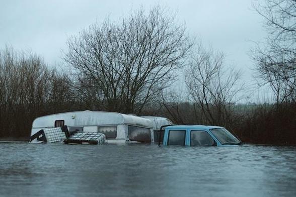 Photo of a flooded scene. Gloomy trees stand in the background as a truck, trailer, and toppled water tanks are half-submerged.
