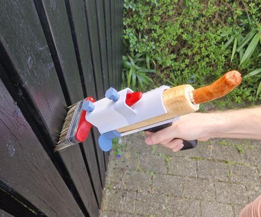 photo of a 3d printed gadget, attaching a large paint brush to a hotdog, held in a pistol grip. Paint brush is held against a black fence and in the background is a hedge.