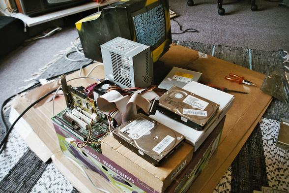 Old PC without a case, on a audergine-box. you see a motherboard with isa and pci slows, a controller sticking out unused, some harddisks on cardboard boxes attached with flat-ribbon-cables, a power supply lies next to it with a cdrom drive somewhere and a spray-painted CRT monitor showing white on black text. All of this on a carpet on the floor. We see in a corner a old tv and keyboard hiding behind the monitor.