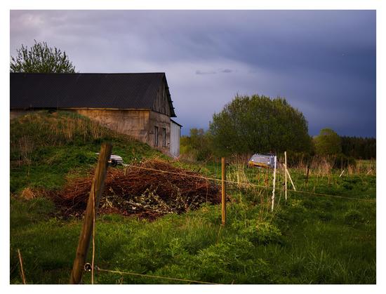 En mörk, liggande bild. I förgrunden går ett staket av stolpar med el-stängsel bort mot höger i grönt gräs. I mitten till vänster står en ekonomibyggnad i brun sten och svart tak. Till häger om den en gammal förfallen Mercedes. I bakgrunden gröna träd och en regntung himmel där molnen skiftar från ljust till mörkt stålblått.