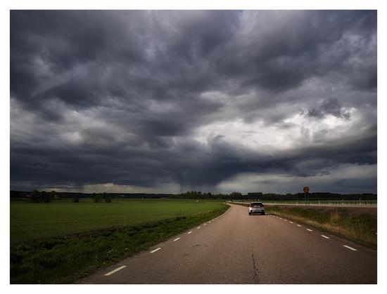 En liggande bild på en silverfärgad bil på väg bort. Till vänster ett grönt fält, till höger ett dike med en större väg på andra sidan. I bakgrunden en dramatisk himmel med mörka moln och mitten, långt bort regn.