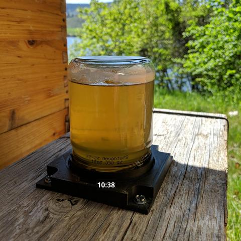 A photo showing a small jar used as a feeder for honey bees. The photo has the time taken added and it says 10:38. It is on a 5 frame nucleus hive. There is a 3d printed fixture to hold the 3d printed jar lid that has a screen in it. The jar has 250 grams of honey mixed with 250 grams of water. The honey is from goldenrod flowers and was solid and white. It was harvested from the hives in October 2024.