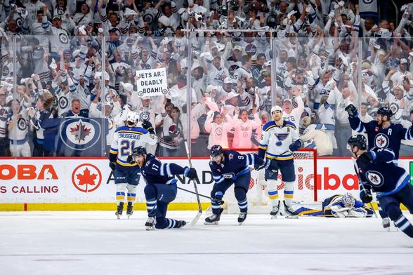 Adam Lowry celebrating on ice in a Jets uniform. You can see all the Winnipeg Jets fans in the background dressed in white and cheering.