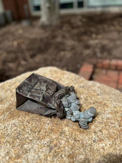 Bronze art of a small paper bag lying on its side, set onto a real rock. Bronzed discs spill out of it. If you look very closely you can see that they’re not coins, but plain wafers with the old Necco logo stamped on them.
