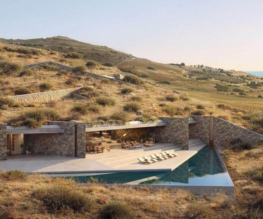 Modern stone house with a pool, nestled on a grassy hillside, surrounded by dry vegetation and rolling landscapes under a clear sky.