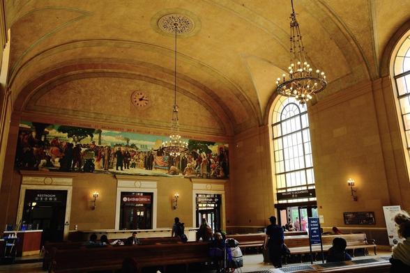 A grand hall features a high arched ceiling with chandeliers, large arched windows, a mural above an entrance, and people sitting on benches.