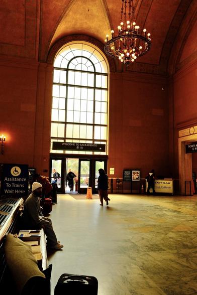 A spacious train station waiting area features benches, travelers, large arched windows, and an elegant chandelier.