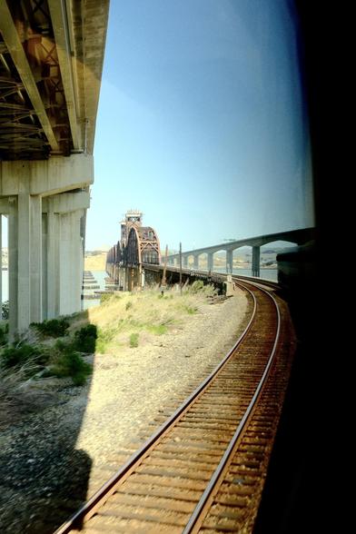 Railroad tracks curve under a large bridge beside a rusted structure against a clear blue sky.