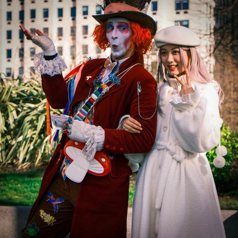A young woman dressed in a white wool coat with heart-shaped silver buttons, a white jumper with a high polo neck, and an off-white  cap is holding her left hand up in a V-sign, standing arms linked to the right of a street performer dressed as the Mad Hatter from the books Alice's Adventures in Wonderland and Alice Through the Looking Glass. He is dressed in a long plum-red coat open at the front, with ten cotton reels lined vertically down his left lapel and a large red and white mushroom pinned lower down; similar coloured trousers with logos down the right leg featuring a peacock, a pink and blue bird, some flowers, and a green leaf; a white shirt with three overlaid bow ties in brown, pink, and blue; various coloured ribbons pinned to his right shoulder; and a black top hat with a pink band. His hair is bright red and his face is painted white with pink and purple makeup. His lacy shirtsleeves protrude from his coat cuffs and he is wearing lace mittens and holding a pink and white tea cup in his left hand. His right hand is held up past his shoulder with open palm facing upwards.