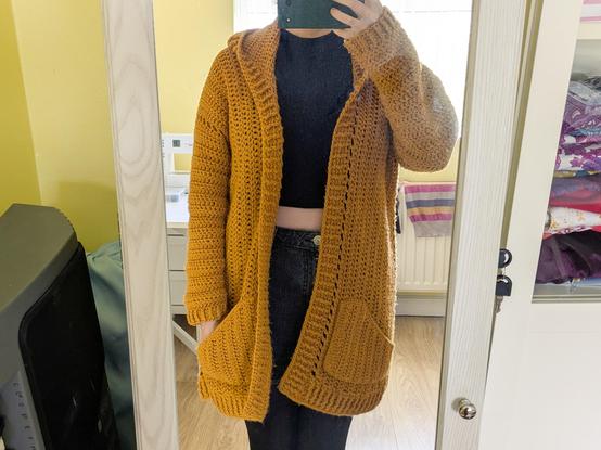 Cropped mirror selfie that cuts off my head and the bottom half of my legs. I'm wearing an indistinct all-black outfit, but over the top of it all is a long golden yellow crocheted cardigan with chonky ribbing, a hood, long sleeves and of course pockets. If you zoomed in you would see many bobbles and fuzzy bits and general signs of wear.