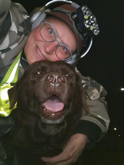 Night time selfie of a person and a chocolate brown Labrador with greying snout. The person is wearing a white-grey military camouflage jacket, yellow safety harness, red cap, silver headphones and blue-rimmed glasses. On their cap rests a headstrap torch in off position. Their free hand is around the dog's shoulders. Both are facing the camera. The dog's mouth is slightly open, showing off some blep. The person's head is resting on the dog's head. They're smiling, grateful for their time together.