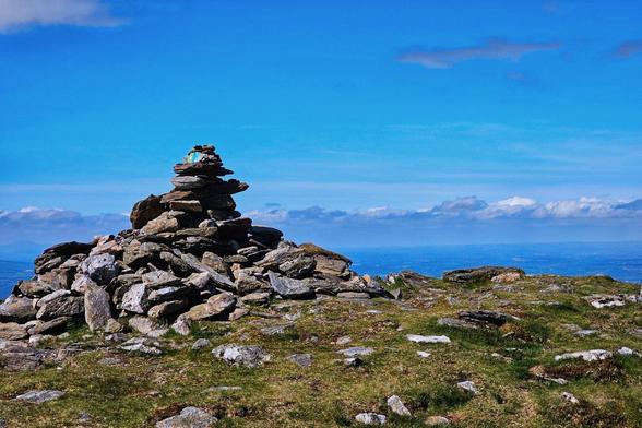 A landscape photo showing a rocky cairn on a grassy hilltop, with a blue sky and scattered clouds in the background. A distant view of the sea is visible on the horizon.