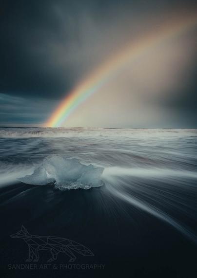 A striking long-exposure photo captures a luminous chunk of glacier ice resting on the black volcanic sands of Diamond Beach in Iceland. The incoming tide blurs into silky streaks around the ice, creating a dynamic sense of motion. Above the churning sea, a vivid rainbow arcs across a moody sky, casting a serene contrast to the stormy ocean scene.