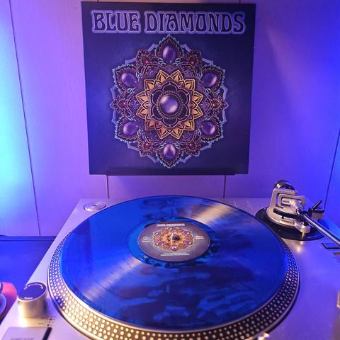 A blue vinyl record sits on a turntable. Behind the turntable, a vinyl album outer sleeve is displayed. The front cover shows an intricate design featuring that looks like a large brooch with multiple gems and floral patterns.