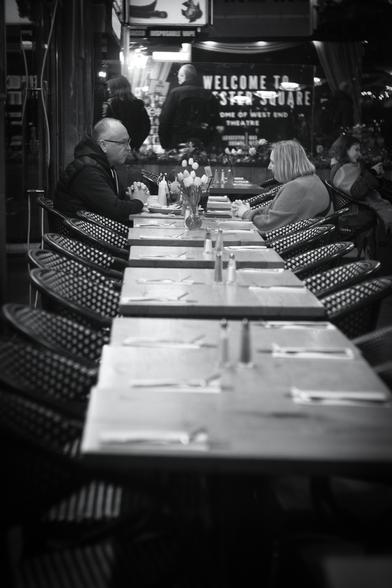 Black and white. The al fresco dining area of a central London cafe/restaurant. Eleven tables are in a row, each with a wicker armchair, seen from the near end, all with knife and fork, some with salt and pepper. A late middle-aged couple are sitting on opposite sides of the ninth table, the man on the left and the woman on the right, both with their hands clasped and wrists on the table, mirroring each other. He has a plate of food in front of him, she has her phone on the table. Both have a tall glass of drink with a straw. There is a vase of nine tulips straddling the seventh and eighth tables. Beyond them are a few more similar tables and chairs, with a young woman sitting at one of them. Beyond those is the window of the cafe through which can be seen the letters, "Welcome to Leicester Square, home of West End theatre". A man and a woman are standing inside the cafe.