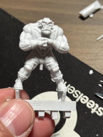 A mini of an orc in a wrestling uniform in gray plastic. It is still on the spruce. Between it’s legs is a little piece of spruce looking a bit inappropriate.