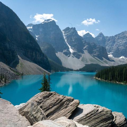A photograph looking over a rock towards a pristine blue lake surrounded by mountains.