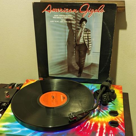 Un tocadiscos de vinilo Teac con una alfombrilla de diseño tie-dye reproduce el disco de vinilo de la banda sonora original de la película "American Gigolo". La portada del álbum muestra a Richard Gere, el protagonista de la película, vestido con un traje elegante. La imagen está tomada desde una perspectiva ligeramente elevada, mostrando tanto el tocadiscos como la portada del álbum con claridad.