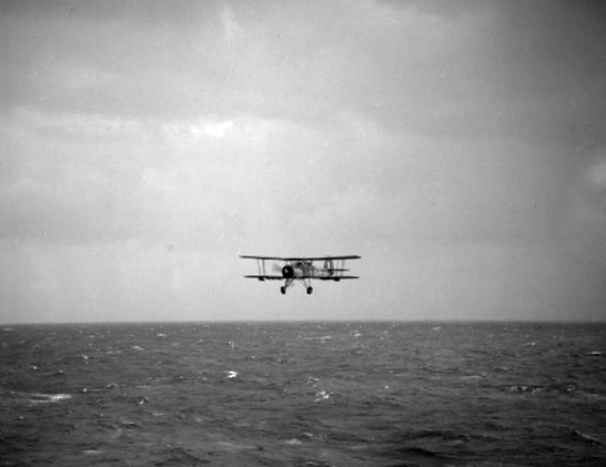 A black and white photo of a biplane (Fairey Swordfish) flying low above the water after the attack on the Bizmarck.  There is no longer a torpedo hanging underneath the plane.