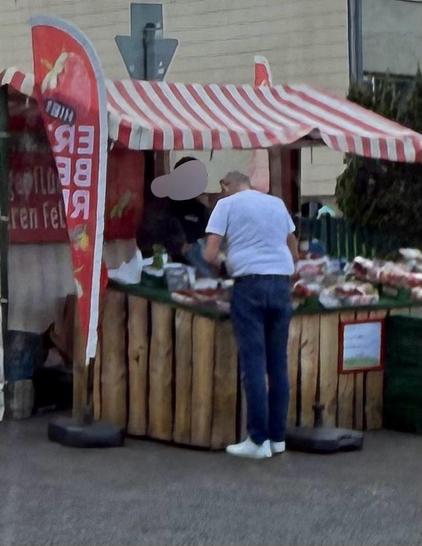 Ein Marktstand mit einem rot-weiß gestreiften Vordach, der Produkte verkauft. Ein Kunde in weißem Hemd und blauer Jeans unterhält sich mit einem Verkäufer hinter dem Stand, der mit verschiedenen Waren, wahrscheinlich Obst oder Gemüse, bestückt ist.