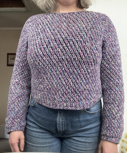Photo of a headless me wearing jeans and a purple crocheted weather. The yarn is mainly purple with some flecks of green, yellow and blue. The crochet pattern makes it look like there are diagonal stripes. The sweater is well fitting and short, stopping just on the waist of my jeans.