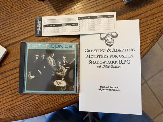 The image features a CD case for "Here Are The Sonics," a black and white photo of the band on the cover. Next to it is a booklet titled "Creating & Adapting Monsters for use in Shadowdark RPG" by Michael Putlack. Also there is a bookmark with quick Shadowdark monster stats.
