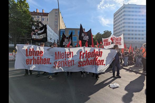 Das Foto von © Sabine Scheffer zeigt das  Fronttransparent mit der Losung „Löhne erhöhen, Mieten senken, Frieden schaffen“ und dahinter laufende Menschen. Im HIntergrund sind weitere Transparente zu sehen.