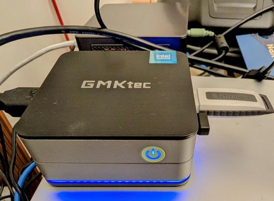 A compact GMKtec mini PC is shown powered on, with a glowing blue LED ring around the power button and base. It has multiple connected cables and a Kingston USB flash drive plugged into the side. The device sits on top of another piece of equipment surrounded by various tech accessories and cables.