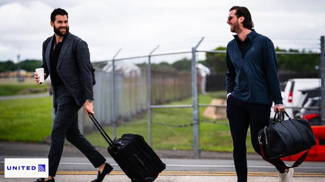 Tom Wilson and Logan Thompson smile while holding their luggage.