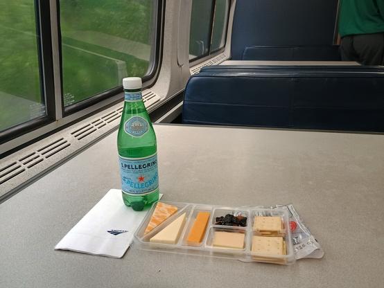 A photo of a prepackaged cheese tray, a bottle of Pellegrino, and an Amtrak logo napkin on a grey lounge car table with dark blue banquette seats, with blurry green trees passing outside the window.