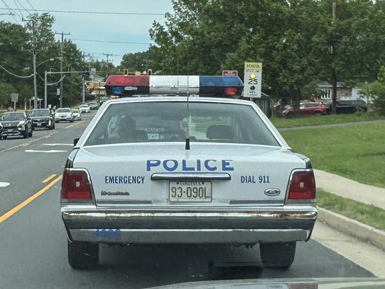 The image shows a white police car with a red and blue emergency light bar on top, positioned on a road. The car is a Ford, as indicated by the badge on the rear. The license plate reads "93-0901." The word "POLICE" is prominently displayed in blue letters on the back of the car, along with the words "EMERGENCY" and "DIAL 911." The car is driving on a road with a yellow dividing line, and there are other vehicles visible in the background. A speed limit sign indicating "SCHOOL 25" is visible on the right side of the image. The environment appears to be a suburban area with trees and houses in the background.

Provided by @altbot@fuzzies.wtf, generated privately and locally using Ovis2-8B

🌱 Energy used: 0.169 Wh