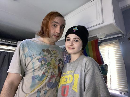A person with reddish-brown hair wearing a graphic t-shirt featuring anime characters stands next to a person in a black beanie and oversized gray shirt. They are in a room with light from a window and a rainbow flag in the background.