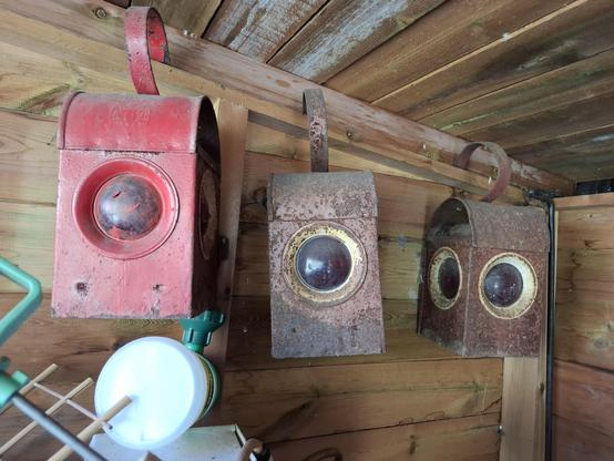 Three red (rusty in two cases) parrafin lamps. They are cuboid with doomed tops and a hanging handle. Each of the four sides has a red glass porthole window.