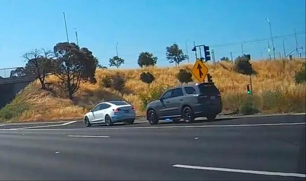 A gray SUV pulling over a white Tesla on the side of the highway.